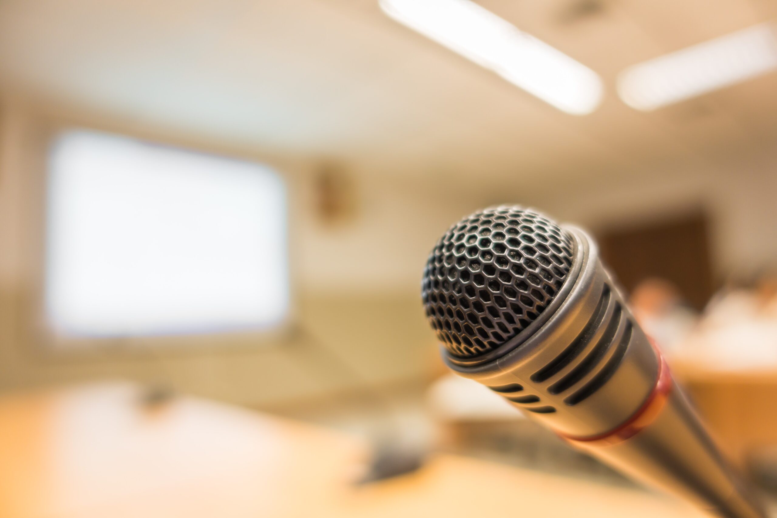 Photograph of a microphone infront of a conference room. The microphone is close to the camera, with the background heavily blurred. Overall a warm and inviting atmosphere.